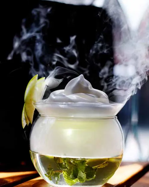 A glass bowl cocktail topped with white foam and apple slices, with dry-ice vapour rising against black