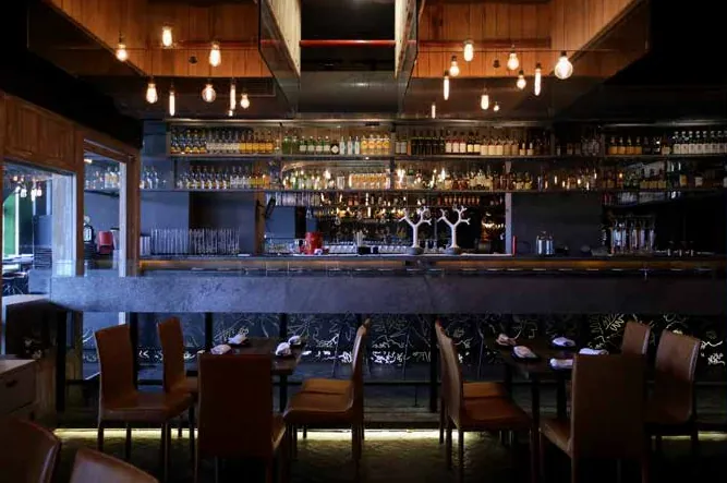 Dimly lit bar counter with blue-grey back bar, pendant bulbs and rows of chairs