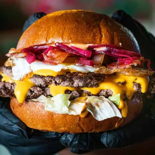Gloved hands holding a double cheeseburger with fried egg, pickled onion and lettuce