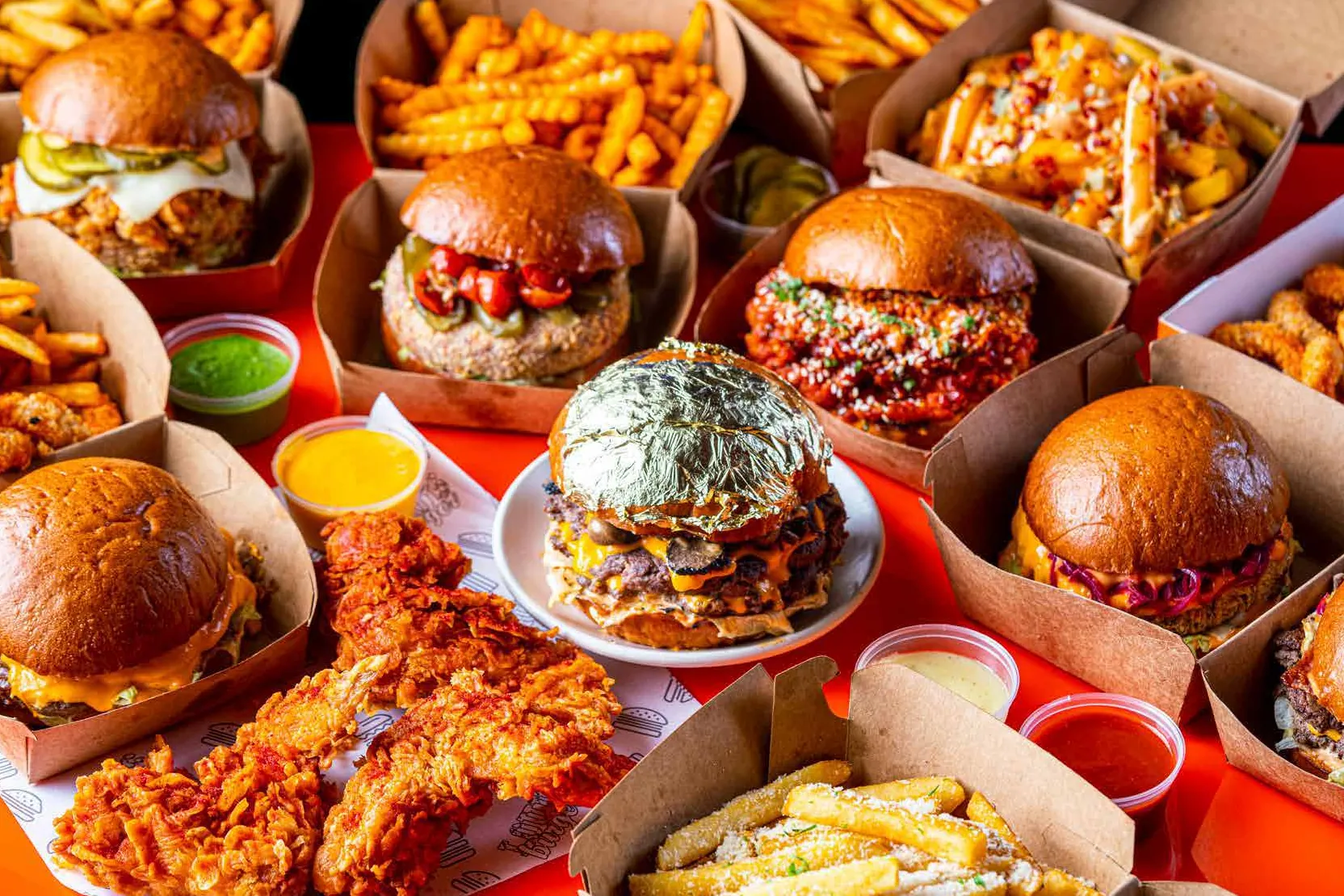 A spread of Louis Burger burgers, loaded fries, fried chicken and dips in kraft boxes on a red table, a gold-leaf burger at centre