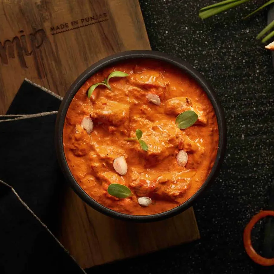 Butter chicken in a black bowl garnished with almonds and herbs, on a wooden board with naan