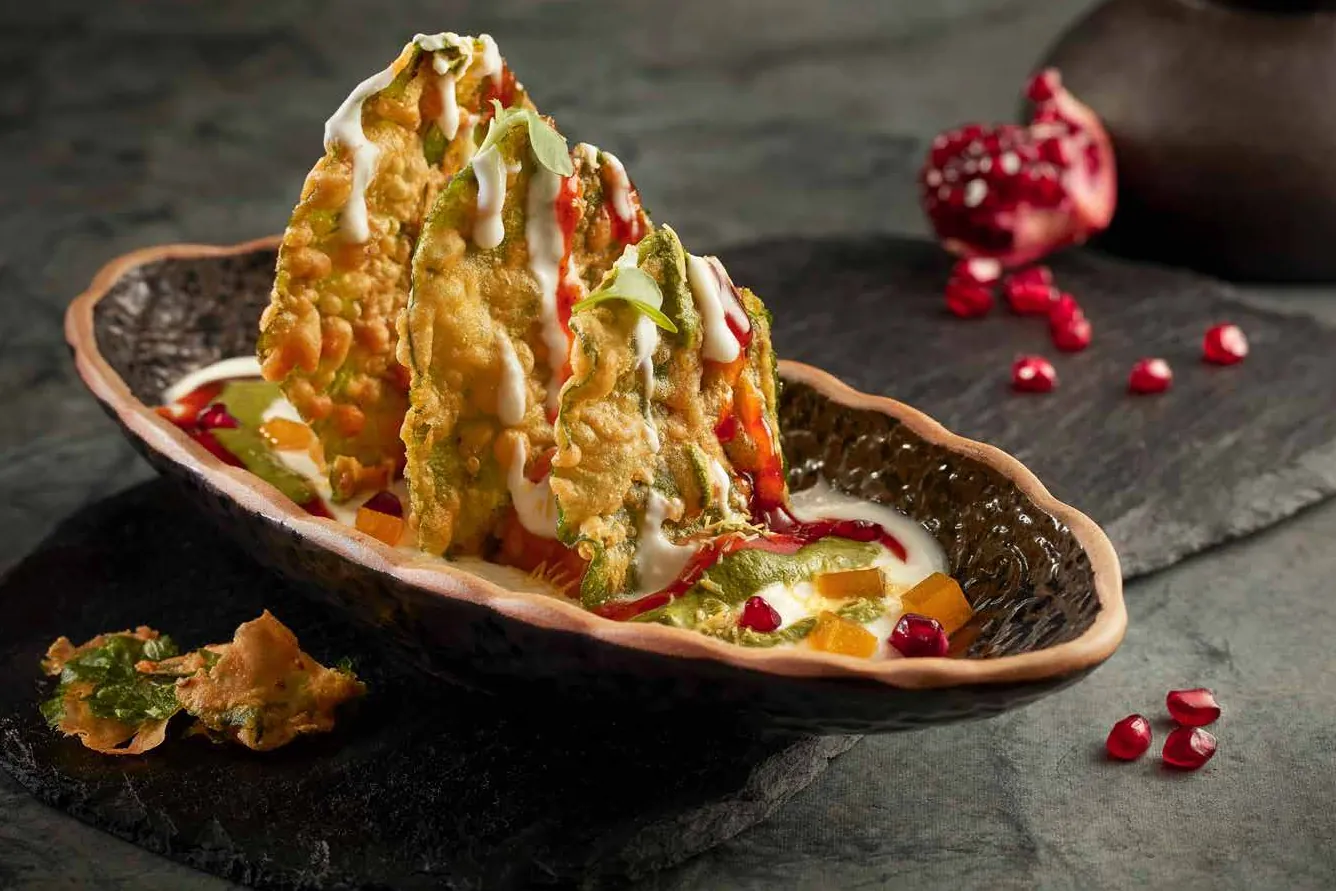 Crisp spinach-leaf fritters drizzled with yoghurt and chutney in a clay dish on dark slate, scattered with pomegranate seeds