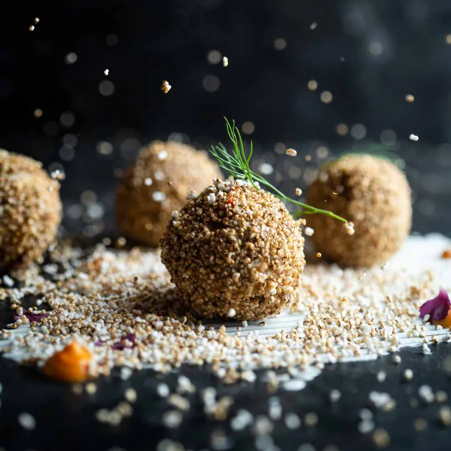 Amaranth-crusted spheres on a dark surface scattered with grains, the front one topped with a sprig of dill