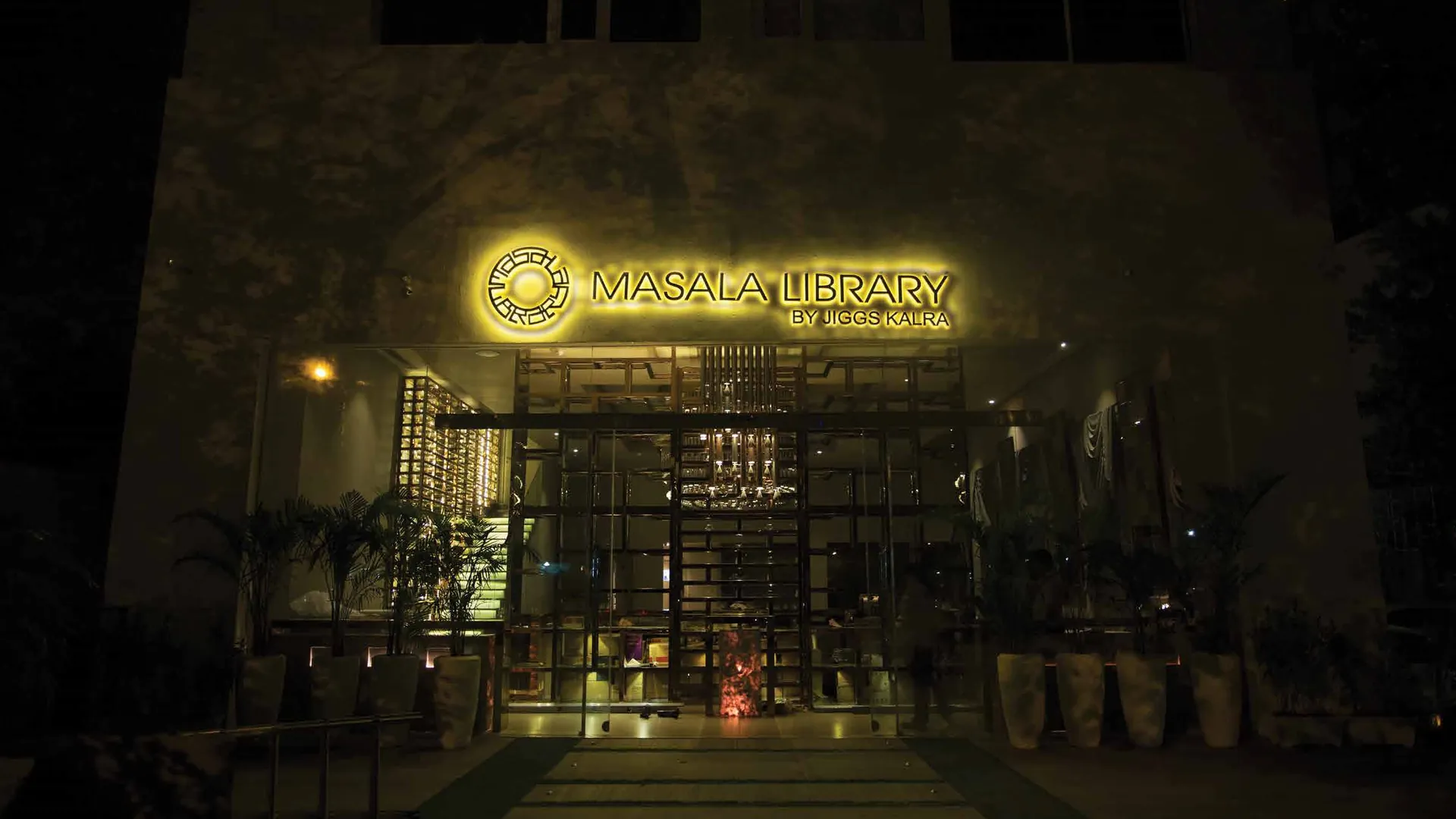 Masala Library by Jiggs Kalra at night, the illuminated gold logo sign above a glass-and-lattice entrance flanked by potted palms