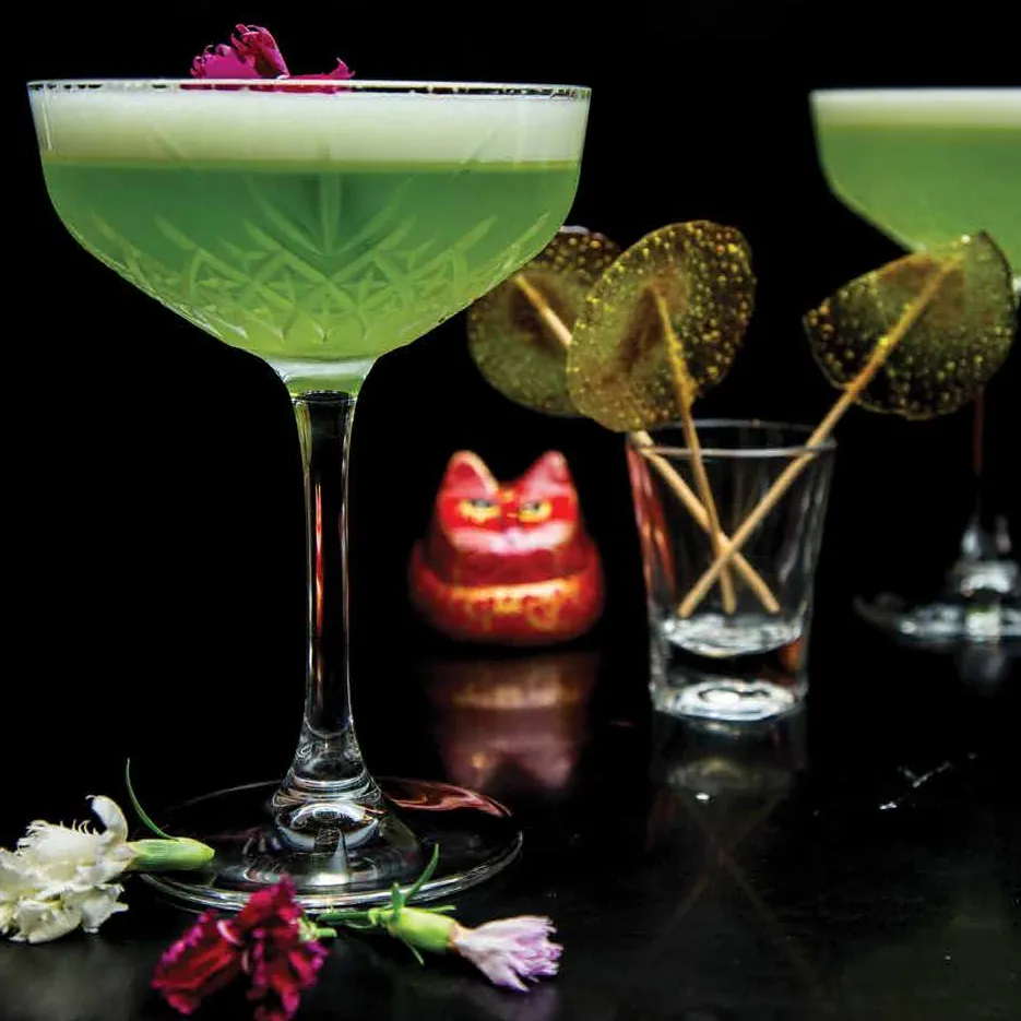 Bright green foamy sours in cut-crystal coupes beside a red lucky-cat figurine, against black