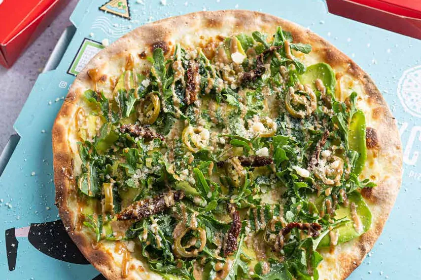 Rocket and olive topped pizza on a light-blue Slyce box beside red Slyce Goodies boxes