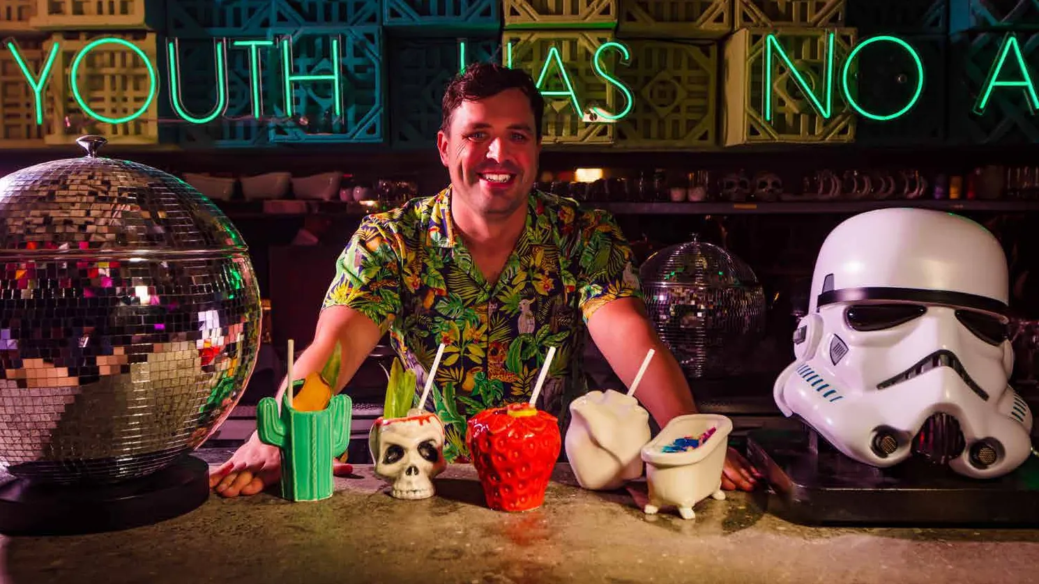 Smiling bartender in a tropical shirt behind the YOUnion bar with novelty tiki cocktails, a disco ball and green neon lettering